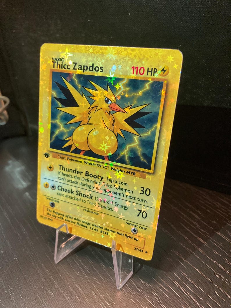 Puede incluir: Una tarjeta coleccionable hologr&aacute;fica con "Thicc Zapdos" con una ilustraci&oacute;n amarilla y azul. La tarjeta muestra texto que incluye "Thunder Booty" y "Cheek Shock" con 110 HP. La tarjeta est&aacute; apoyada en un soporte de pl&aacute;stico transparente.