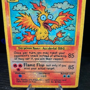 May include: A custom-made trading card featuring a cartoonish, yellow and orange bird character named "Derptres." The card has a humorous design with the text "Derpemon Power: Accidental BBQ" and "Flame Flop." The card is displayed on a clear stand.
