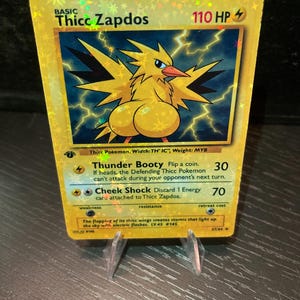 Puede incluir: Una tarjeta coleccionable hologr&aacute;fica con un p&aacute;jaro de dibujos animados amarillo con el texto "Thicc Zapdos". La tarjeta tiene un fondo azul con rayos y el texto "Thunder Booty" y "Cheek Shock". La tarjeta est&aacute; apoyada en un soporte transparente.