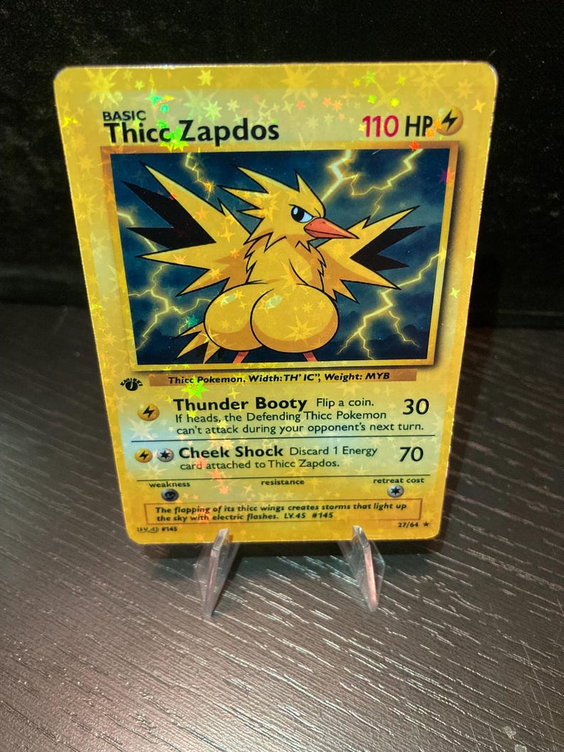 Puede incluir: Una tarjeta coleccionable hologr&aacute;fica con un p&aacute;jaro de dibujos animados amarillo con el texto "Thicc Zapdos". La tarjeta tiene un fondo azul con rayos y el texto "Thunder Booty" y "Cheek Shock". La tarjeta est&aacute; apoyada en un soporte transparente.