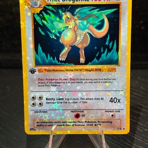 May include: A holographic Thicc Dragonite Pokémon card. The card features a cartoon dragon with a large posterior, in shades of orange, blue, and green. The card has a gold border and includes text such as "Thicc Dragonite 100 HP".