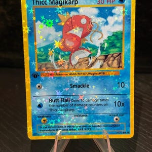 Może przedstawiać: Holograficzna karta kolekcjonerska z "Thicc Magikarp" i 30 HP. Karta ma niebieskie tło z czerwoną ilustracją Magikarpa. Tekst: "Smackle" i "Butt Flail". Karta jest prezentowana na przezroczystym stojaku.