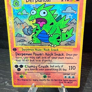 May include: A holographic Derpantar Pokémon card, featuring a green dinosaur-like creature with a rock. The card has text including "Stage 2," "140 HP," and "Clumsy Crush." The card is displayed on a clear stand.