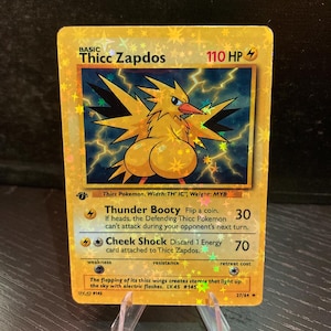 Puede incluir: Una tarjeta coleccionable hologr&aacute;fica dorada con un personaje de p&aacute;jaro de dibujos animados llamado "Thicc Zapdos". La tarjeta tiene texto que incluye "Thunder Booty" y "Cheek Shock". La tarjeta se muestra en un soporte de pl&aacute;stico transparente.