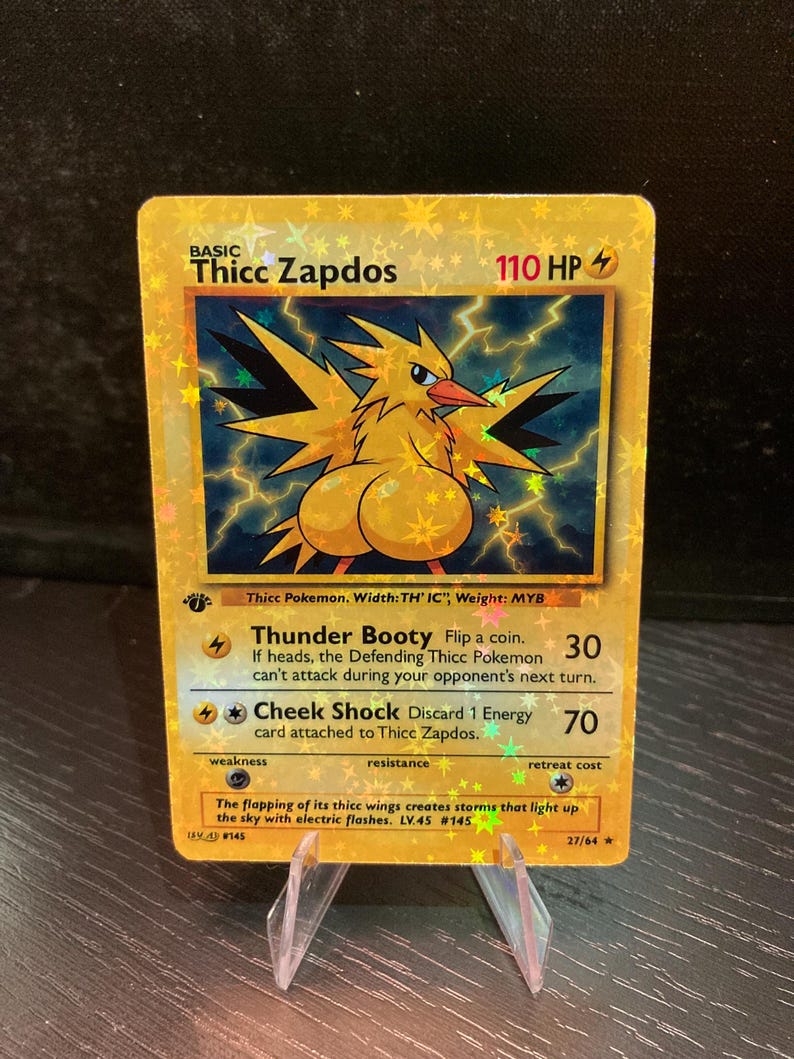 Puede incluir: Una tarjeta coleccionable hologr&aacute;fica dorada con un personaje de p&aacute;jaro de dibujos animados llamado "Thicc Zapdos". La tarjeta tiene texto que incluye "Thunder Booty" y "Cheek Shock". La tarjeta se muestra en un soporte de pl&aacute;stico transparente.