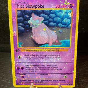 Puede incluir: Carta coleccionable Pokémon Thicc Slowpoke con borde morado y diseño holográfico. La carta presenta una ilustración de un Slowpoke rosa con el texto "Thicc Slowpoke" y "50 HP". La carta se muestra en un soporte transparente.