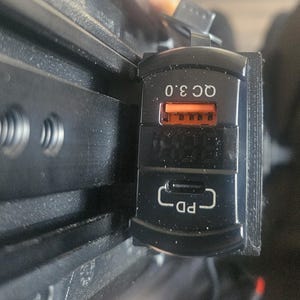 May include: A black rectangular dual-port USB charger featuring an orange USB-A port labeled QC 3.0 and a USB-C port labeled PD. The device has a digital display screen positioned between the two ports and is mounted on a black plastic rail system.