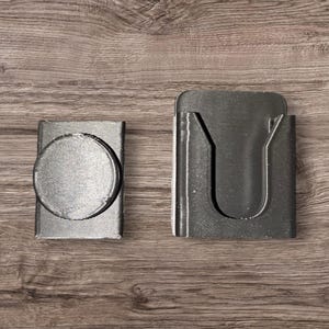 May include: Two small, silver-colored metal objects. One is rectangular with a circular knob on the front. The other is a rectangular holder with a U-shaped opening. Both objects are on a wooden surface.