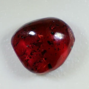 Puede incluir: Una piedra preciosa pulida de color rojo intenso con una forma suave, redondeada e irregular. La piedra presenta una calidad translúcida con inclusiones oscuras visibles en el interior y una superficie brillante que refleja la luz.