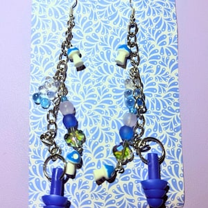 May include: A pair of handmade earrings featuring a whimsical design. Each earring has a silver chain, blue and white mushroom charms, and blue earplugs. The earrings also include blue and clear beads, and are displayed on a blue patterned card.