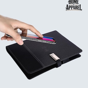 May include: A black rectangular portfolio case with a textured fabric cover, a smooth black leather-like side panel, and a metallic silver clasp closure. The case features a wireless charging icon on the front cover and the text HOME OF APPAREL.