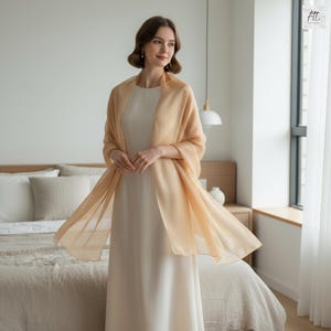 May include: A peach-coloured shawl draped over a cream-coloured dress. The shawl is made of a lightweight, flowing fabric. The woman is standing in front of a bed with neutral-toned bedding. The room has a minimalist design with natural light.