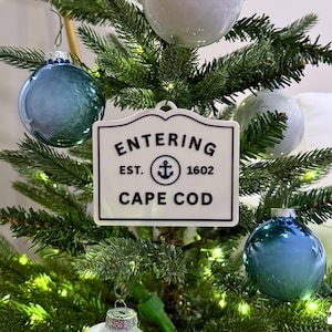 Puede incluir: Un árbol de Navidad decorado con adornos azules y blancos y un letrero blanco que dice "ENTERING CAPE COD EST. 1602" con un símbolo de ancla. El árbol tiene agujas verdes y pequeñas luces.