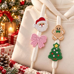 May include: A cream-coloured hoodie featuring two white drawstrings decorated with embroidered patches. The left string has a Santa Claus face and a pink bow. The right string features a gingerbread man and a green Christmas tree with a star.