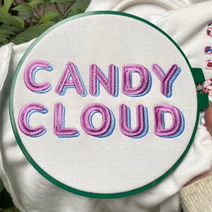 May include: Embroidered hoop art with the words "CANDY CLOUD" in pink, outlined in light blue, on a white fabric. The embroidery is set in a green hoop. The design is on a white background.