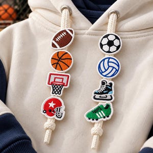May include: A cream-colored hoodie with navy blue sleeves featuring two thick cream drawstrings. Each drawstring is adorned with a vertical row of embroidered sports-themed patches, including a football, basketball, hoop, helmet, soccer ball, volleyball, ice skate, and cleat.