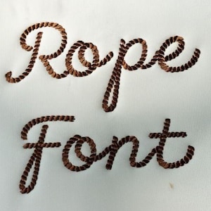 May include: The words "Rope font" are embroidered in a brown rope-like design on a white background. The letters are stylized to resemble rope, creating a nautical or rustic aesthetic. The font is used for decorative purposes.
