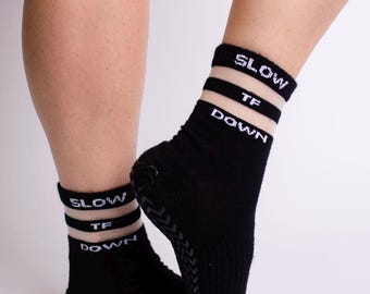 May include: Black socks with the words "SLOW TF DOWN" in white lettering. The socks have a white stripe and a textured sole. The socks are worn on a person's feet.
