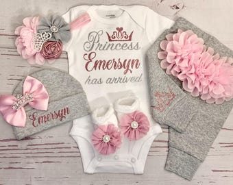 May include: A newborn baby outfit set in white and gray, featuring a onesie with "Princess Emersyn has arrived" text, a matching hat, pants, socks, and floral accents. The set includes a headband and is decorated with pink and silver glitter.