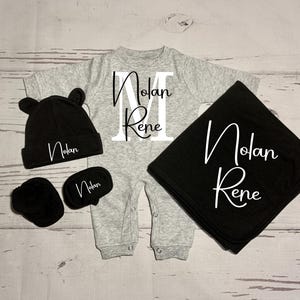 May include: A gray baby onesie with the name "Nolan Rene" and the letter "M" on the front. The set also includes a black hat, a pair of black mittens, and a black blanket with the name "Nolan Rene" embroidered on it.