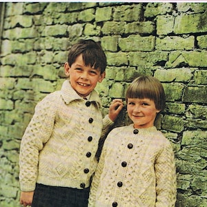 Puede incluir: Dos niños con cárdigans de punto color crema con cierre de botones. El cárdigan de la izquierda tiene un patrón de punto de cable. El cárdigan de la derecha tiene un patrón de punto de diamante. Los niños están de pie frente a una pared de piedra. La imagen incluye el texto "Báinín wool mahony's of blarney 649 10d".