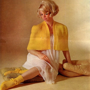 May include: A woman wearing a yellow knitted cape and white nightgown sits on a cushion. She is wearing yellow knitted socks.