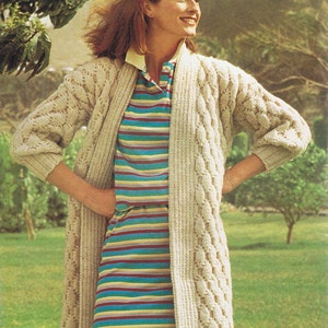 Może przedstawiać: Kobieta w długim, kremowym kardiganie z dzianiny z wzorem warkocza, nałożonym na paskowaną niebiesko-żółto-czerwoną koszulę. Stoi na trawiastym polu z drzewami w tle.