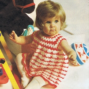 Könnte beinhalten: Ein rot-weiß gehäkeltes Babykleid. Das ärmellose Kleid hat ein sich wiederholendes Muster aus weißen Formen auf rotem Hintergrund. Das Kleid wird von einem Baby getragen, das mit Spielzeug sitzt.
