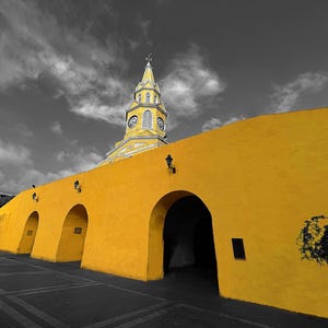 May include: A vibrant yellow wall with arched entryways and a clock tower against a black and white sky. The building has a classic design with a yellow clock tower and a long, yellow wall with multiple arched openings.