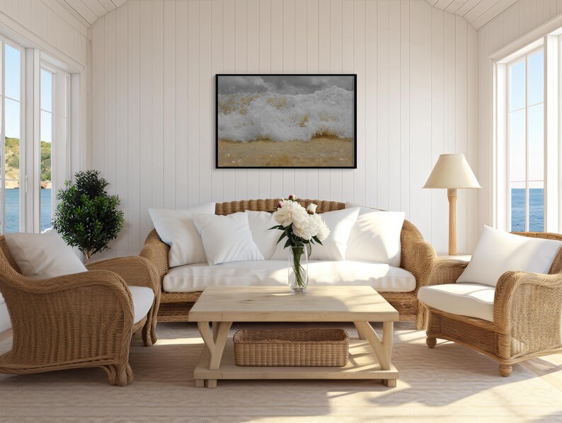 May include: A coastal-themed living room with wicker furniture, including a sofa, two armchairs, and a coffee table. A framed picture of ocean waves hangs above the sofa. White cushions and a vase of white flowers add to the room's bright, airy feel. A small green plant is in the corner.