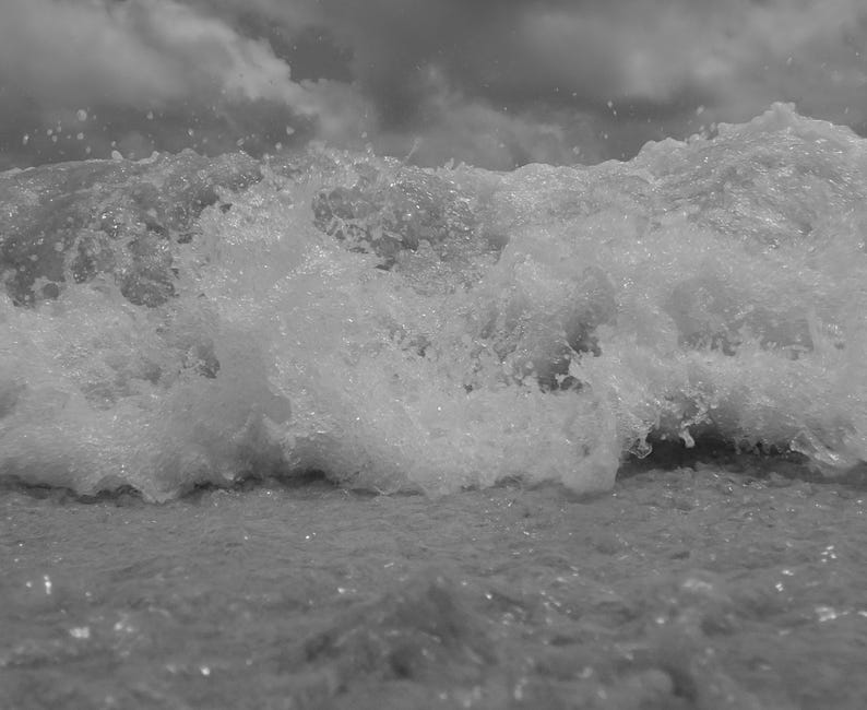May include: A monochrome photograph of a breaking wave. The frothy white water of the wave contrasts with the darker water below and the cloudy sky. The image highlights the power and motion of the sea.