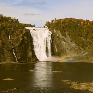 May include: A picturesque landscape showing a large waterfall flowing into a lake. The cliffs are covered in green and autumnal trees. A small rainbow is visible near the base of the falls, creating a scenic view.