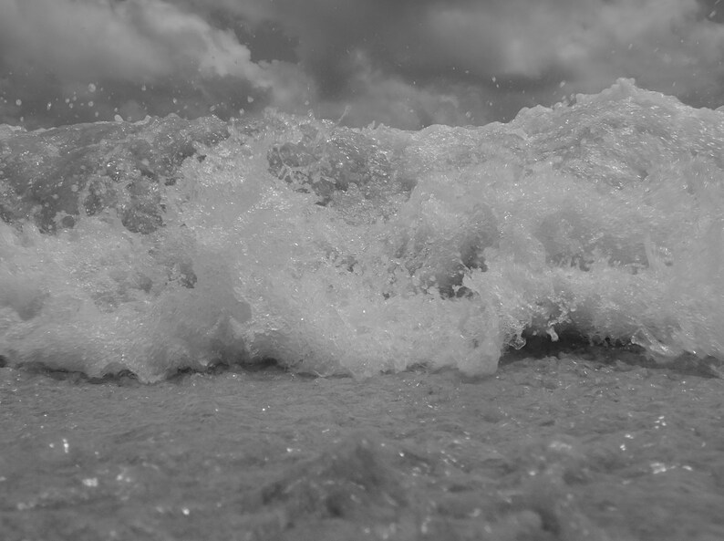 May include: Monochrome image of a breaking wave. The water is frothy and churning, with water droplets suspended. The sky is filled with dark, overcast clouds, creating a stark contrast.