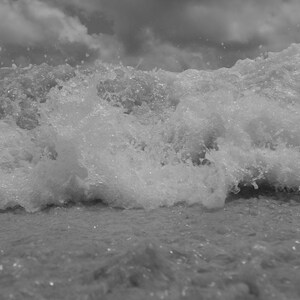 May include: Monochrome image of a breaking wave. The water is frothy and churning, with a cloudy sky overhead. The photograph captures the dynamic motion of the sea.