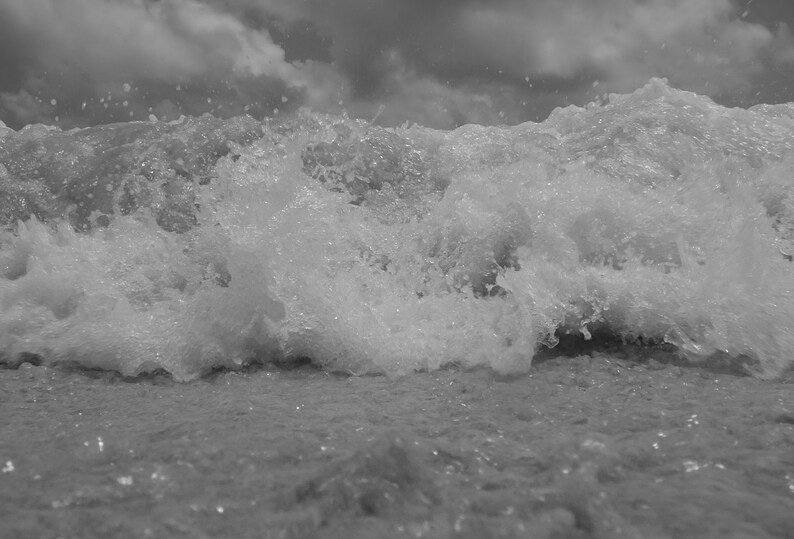May include: Monochrome image of a breaking wave. The water is frothy and churning, with a cloudy sky overhead. The photograph captures the dynamic motion of the sea.