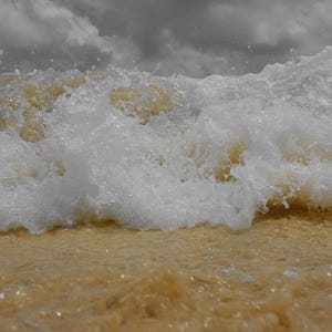 May include: A close-up shot of a wave crashing on a sandy beach. The wave is a mix of white foam and a light brown color, with the sand in the foreground also appearing light brown. The sky is overcast with gray clouds.