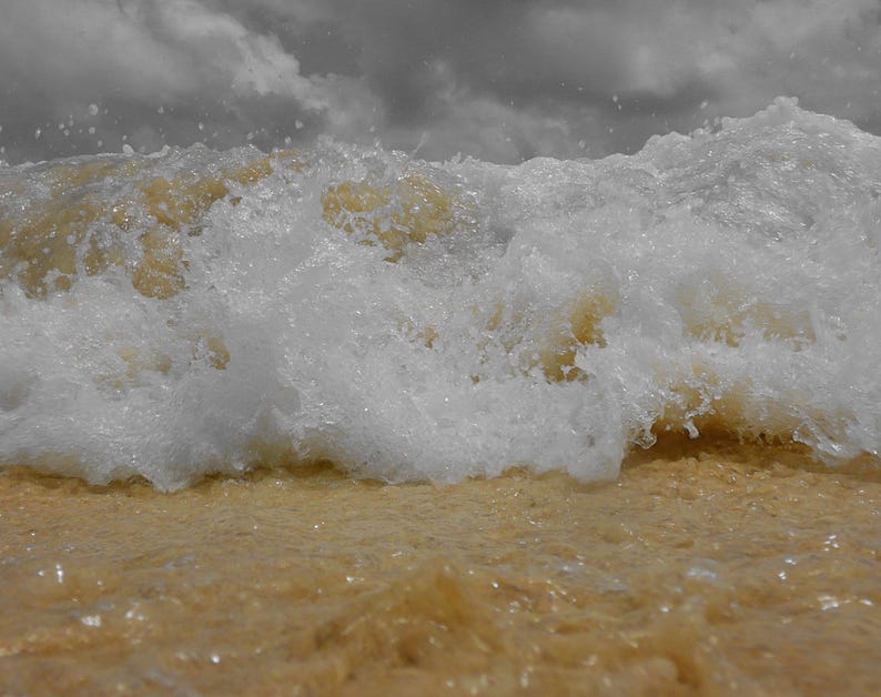 May include: A close-up shot of a wave crashing on a sandy beach. The wave is a mix of white foam and a light brown color, with the sand in the foreground also appearing light brown. The sky is overcast with gray clouds.