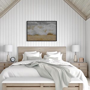 May include: A framed print of a crashing ocean wave hangs above a bed with white bedding. The bedroom has a light wood headboard, matching nightstands, and a white bench at the foot of the bed. Two white lamps sit on the nightstands.