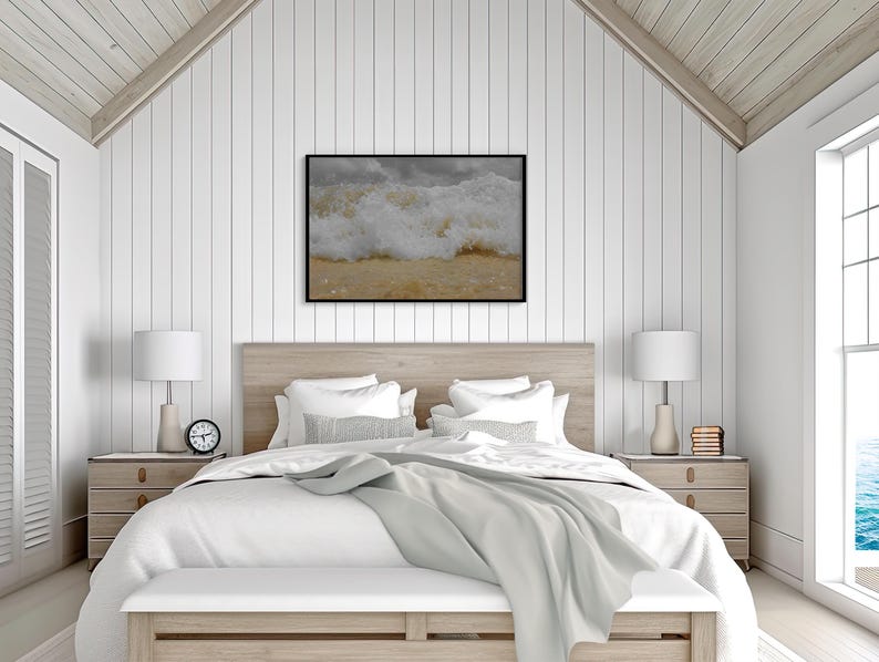 May include: A framed print of a crashing ocean wave hangs above a bed with white bedding. The bedroom has a light wood headboard, matching nightstands, and a white bench at the foot of the bed. Two white lamps sit on the nightstands.