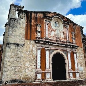 May include: Exterior view of a historic building with a stone facade and an arched entrance. The building features a combination of light-colored stone and reddish-brown stucco. A circular window with ornate detailing is visible near the top, under a partly cloudy blue sky.