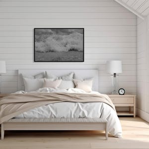 May include: A bedroom featuring a white bed with a beige throw, flanked by two bedside tables with lamps. A black and white framed picture of ocean waves is above the bed. A small clock sits on a bedside table.