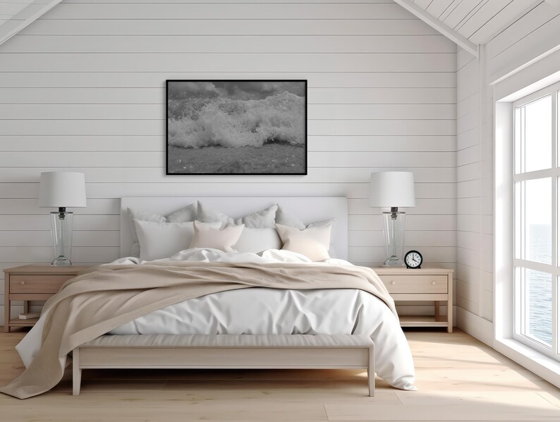 May include: A bedroom featuring a white bed with a beige throw, flanked by two bedside tables with lamps. A black and white framed picture of ocean waves is above the bed. A small clock sits on a bedside table.