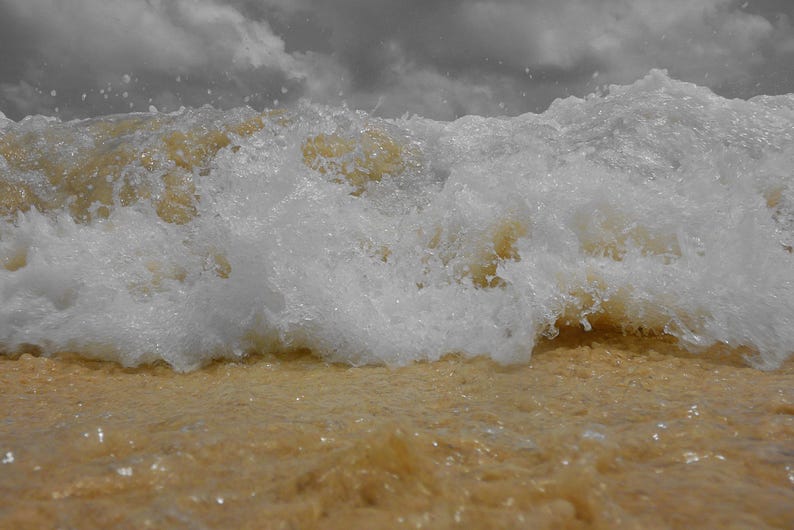 May include: A close-up shot of a wave breaking on a sandy beach. The wave is white and foamy, with a light brown color from the sand. The sky is overcast with gray clouds, creating a dramatic contrast.