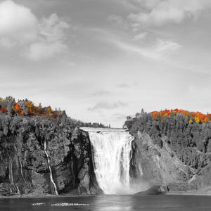 May include: A black and white photograph of a waterfall cascading down a cliff face. The image is mostly grayscale, with the exception of the trees at the top of the cliff, which are in vibrant shades of red, orange, and yellow.