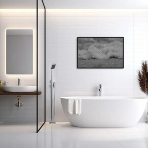 May include: A contemporary bathroom featuring a white, freestanding bathtub, a framed monochrome photograph of ocean waves, and a large illuminated mirror. The room has white tiled walls and a dark wood accent wall.