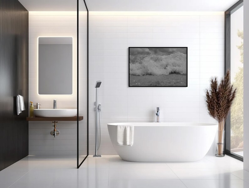 May include: A contemporary bathroom featuring a white, freestanding bathtub, a framed monochrome photograph of ocean waves, and a large illuminated mirror. The room has white tiled walls and a dark wood accent wall.