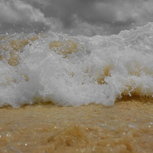Puede incluir: Primer plano de una ola rompiendo en una playa de arena. La ola es blanca y espumosa, con un tinte marrón dorado de la arena. El cielo está cubierto de nubes grises, creando un contraste dramático.