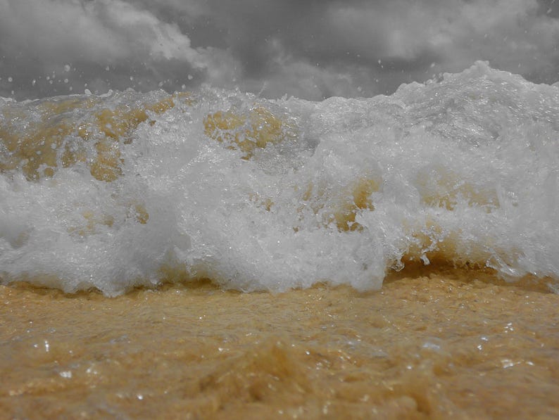 May include: A close-up shot of a wave breaking on a sandy beach. The wave is white and foamy, with a golden-brown hue from the sand. The sky is overcast with gray clouds, creating a dramatic contrast.