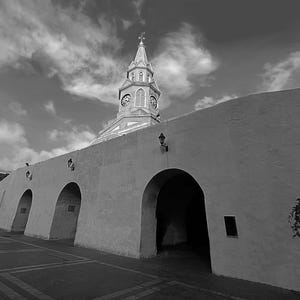 May include: Black and white photograph of a historic building with a clock tower. The building has a white facade with arched entryways and a tall clock tower with a spire. The sky is filled with clouds.