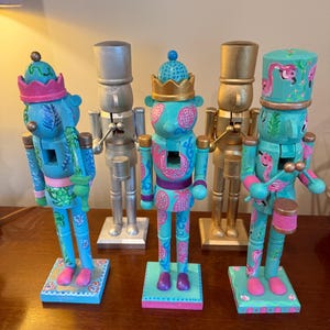 Könnte beinhalten: Fünf dekorative Nussknacker. Die Nussknacker sind in Blau, Silber und Gold bemalt. Einige haben rosa und türkisfarbene Muster, andere Flamingodesigns. Jeder steht auf einem quadratischen Sockel. Die Höhe beträgt ca. 20 cm.